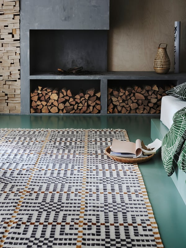 Cozy room with handwoven VAMDRUP rug in beige/brown, soft wool, geometric pattern, under stacked firewood and green-painted low platform with pillows.