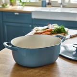 Enamel casserole pan on wooden counter with veggies, designed for even heat distribution.