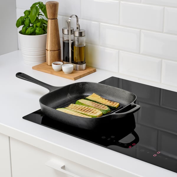 Black cast iron skillet on stovetop with grilled zucchini. Surrounded by seasonings and oil, showing heat distribution and durability.