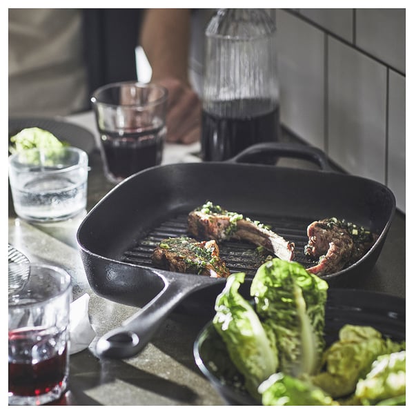 A black cast iron skillet with meat cooking on a kitchen counter, surrounded by glasses of red wine and a bowl of green vegetables.