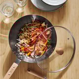 A black carbon steel wok on a wooden table, filled with colorful stir-fried vegetables and noodles, sits beside a glass lid and empty wine glasses.