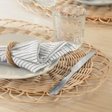 A dining table set with wicker placemats, white plates, folded striped napkins, a knife, and a glass, showcasing a rustic, handwoven design.