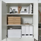 White cabinet with art, design books, and VATTENTRÅG boxes for notes and pens, organized neatly.