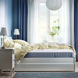 Bright, modern bedroom with blue curtains, large window, white bed with yellow blanket, two lamps, and a large hanging light fixture.