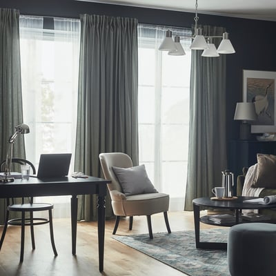 Modern living room with dark walls, large windows, desk, laptop, rug, armchair, pendant lights, coffee table, and curtains.