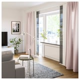 Bright living room with grey couch, coffee table, patterned rug, pink curtains, and large windows showing greenery.