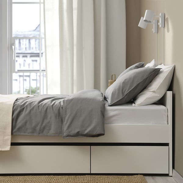 Modern bed with grey linens, storage below, floor lamp, city view.