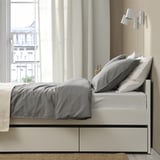 Modern bed with grey linens, storage below, floor lamp, city view.