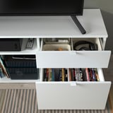 A white VIHALS tv bench with two drawers, one open displaying books, and another with headphones. The top holds a flat-screen tv.