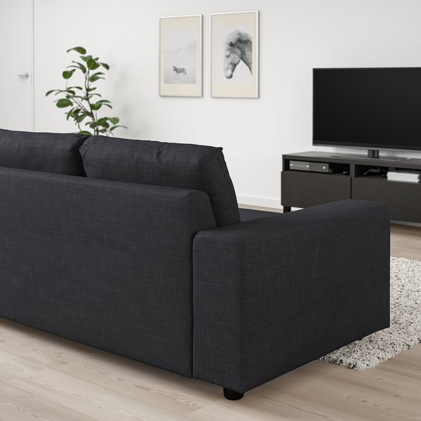 A modern black VIMLE sofa with a chaise longue, positioned in a living room with wooden floors. It is accompanied by a flat-screen tv and a rug.