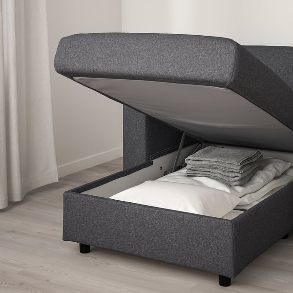 A gray upholstered bed with a hinge-up base revealing stacked blankets and pillows, designed for storage.