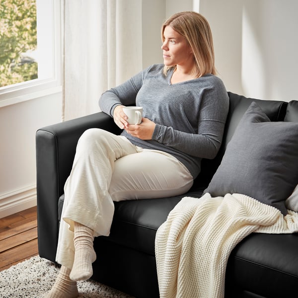 A woman relaxes on a black VIMLE sofa, holding a mug, wearing a gray sweater and cream pants. A beige blanket is draped over the armrest.