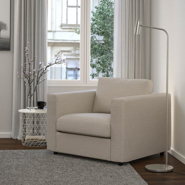 Its a VIMLE beige sofa chair in a minimalist room, adjacent to a tall lamp and small table holding a vase and mug.