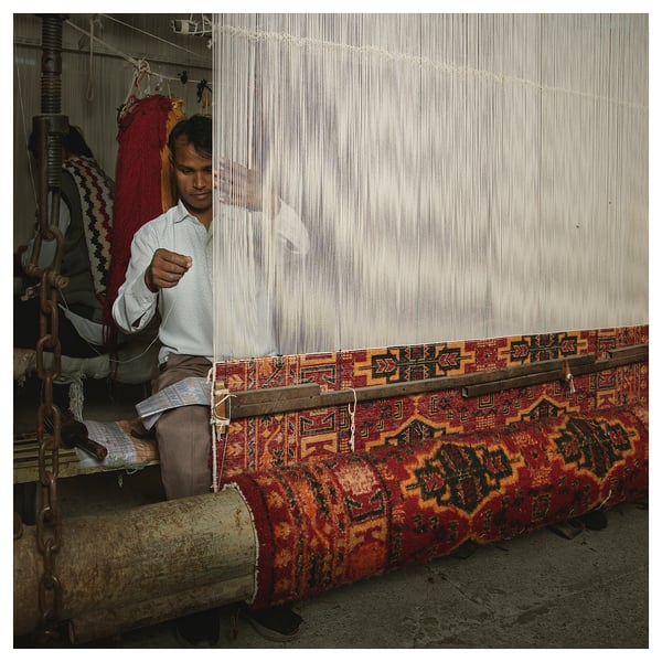 Man weaving colorful, patterned rug on traditional loom. Rug red, yellow, and black designs. Loom has white threads.