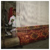 Man weaving colorful, patterned rug on traditional loom. Rug red, yellow, and black designs. Loom has white threads.