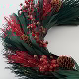 Circular wreath with vibrant red flowers, green foliage, and pinecones, artfully arranged for holiday decor.