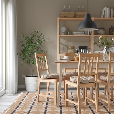 Dining area with wooden chairs & beige cushions, rug, plant, & shelves with decor.