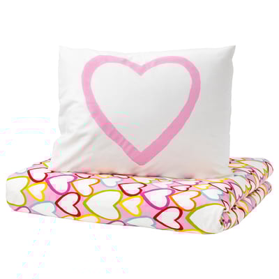 White pillow with pink heart shape on top, resting on colorful heart-patterned pink and white bedsheet.
