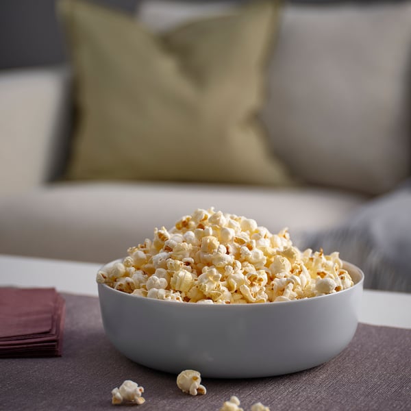 Bowl of popcorn on couch. White VITBLECKA glass bowl, large, round, simple design. Bowl holds significant amount of popcorn, near burgundy napkins.