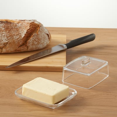 Clear butter dish with lid, holding a pat of butter, beside a cutting board with bread and a knife.