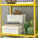 Vibrant yellow shelf with three glass shelves holding storage boxes, books, and decorative cacti.
