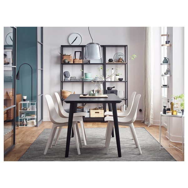 Modern dining space with black table, four white chairs, gray rug, and black-framed glass shelves displaying decor.