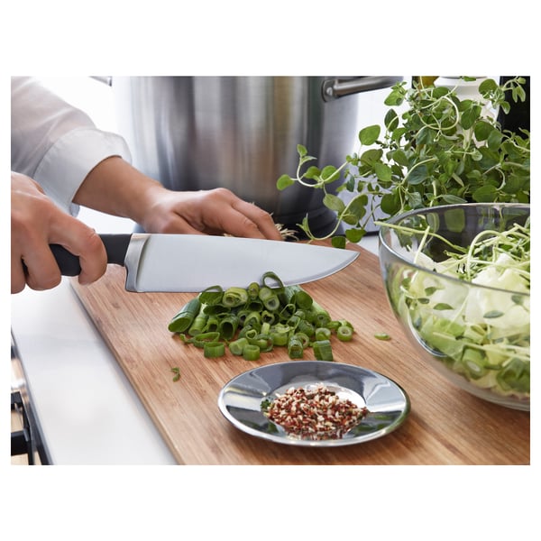 Person chopping scallions on a wooden cutting board with a stainless steel knife. A small plate of chili flakes is nearby.