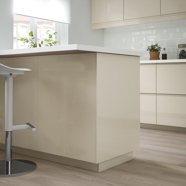 Modern kitchen with high-gloss beige VOXTORP cabinets, sleek handles, and a white countertop. Tall units dominate, with a bar stool to the side.