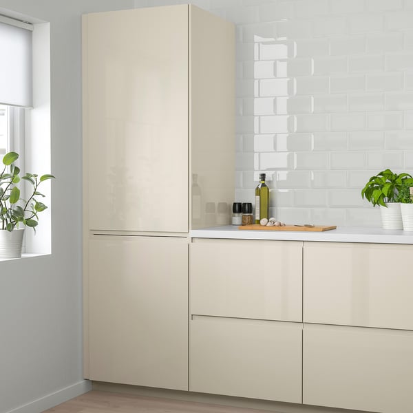 Modern kitchen with large cream-colored VOXTORP cabinets. High-gloss finish. Tall cabinets taller than window. Plants on counter.