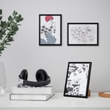 Desk with headset, books, framed art. Wall has two pictures, one on desk. Black frames: floral, woman.