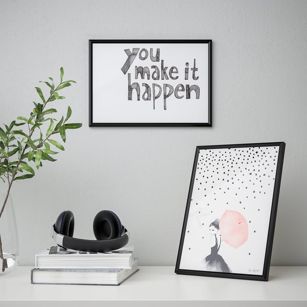 Framed posters on a desk. One says you make it happen, the other shows a woman with a red dot. Headphones on books, plant beside.