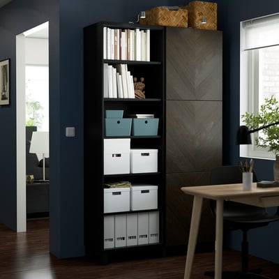 A black BESTÅ storage unit with shelves, housing boxes and books, next to a wooden chair and table. It stands tall against a blue wall.