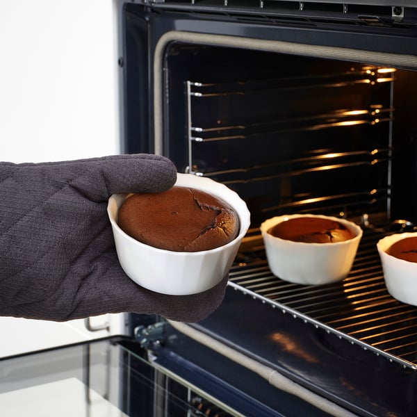 Hand wearing oven mitt holds hot chocolate lava cake in white ceramic dish, being taken out of oven.