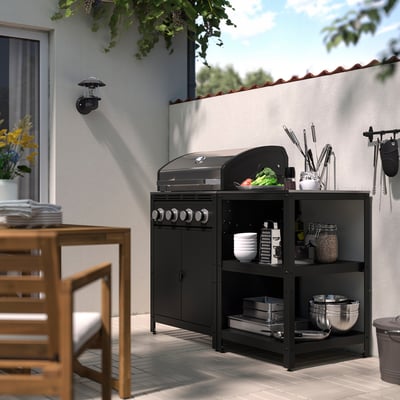 Outdoor kitchen setup with black GRILLSKÄR gas grill and storage shelf, holding plates, bowls, and utensils. Wooden chairs and table nearby.