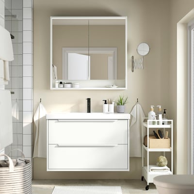 Modern white bathroom vanity with storage. Mirrored cabinet, plant, toiletries, trolley, and basket.