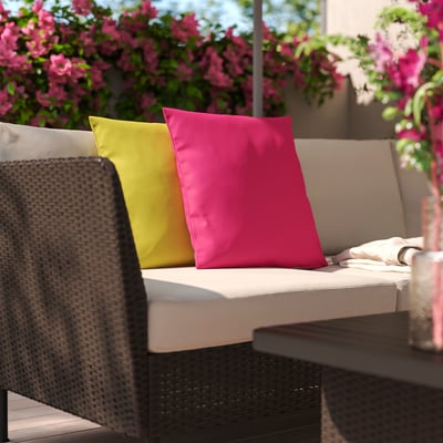 Bright magenta HJÄRTERÖ cushion on outdoor wicker couch, contrasting with large yellow and beige cushions, and surrounded by vibrant pink flowers.