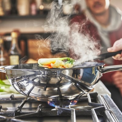A chef cooks in a wok on a gas stove. Steam rises, showing the woks use in stir-frying, demonstrating its non-stick and heat-distributing properties.