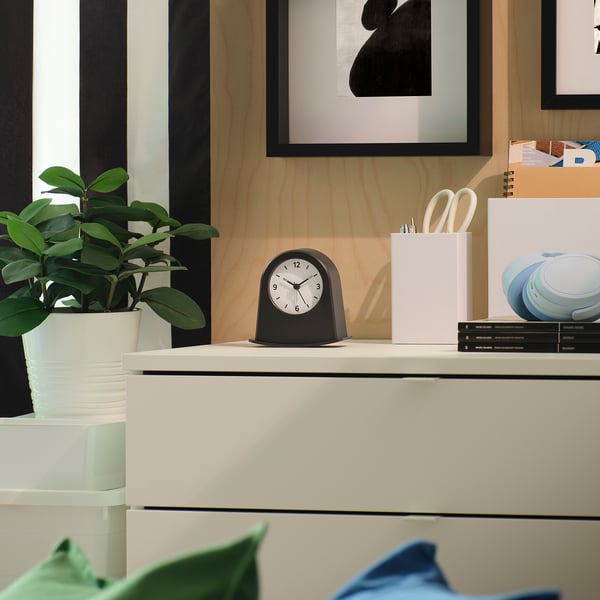 Black KÄRRSÅNGARE alarm clock on desk, apple-sized, with white face. Between plant and pencil holder, cosy workspace with art and wood.