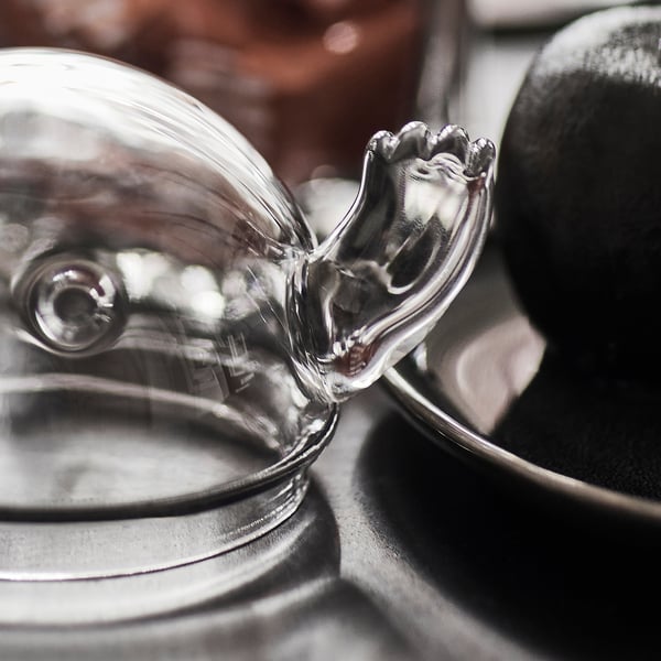 Ghost-shaped glass container with lid; clear, reflective surface; sits on metallic base.