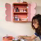 A person organising art supplies in a pink wall cabinet with a shelf, featuring closed doors and scalloped edges, mounted on a light-colored wall.