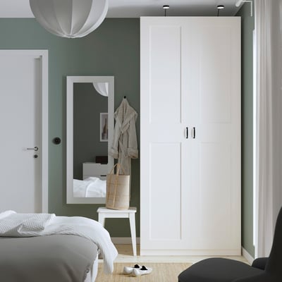 Modern bedroom with white PAX wardrobe, sage walls, black handles, bench, and mirror.