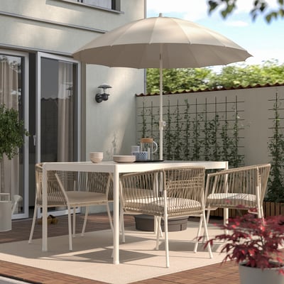 Small patio with white SAMSÖ parasol, wicker chairs, table, and a house with glass doors. Terrace has plants and a fence with greenery.
