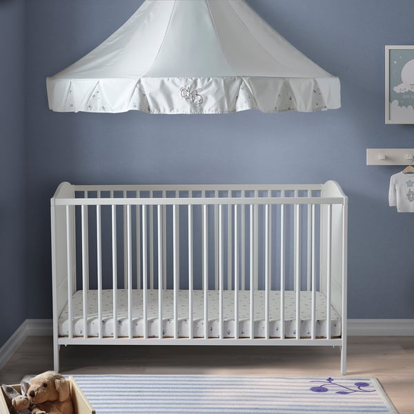 White SMÅGÖRA crib with canopy against blue wall. Spacious, with rounded corners and a rug. Teddy nearby.