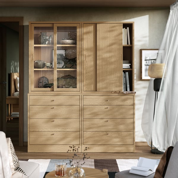 Modern wooden cabinet & drawers set, golden oak, with glass doors & shelves, positioned near a window, taller than a chair.