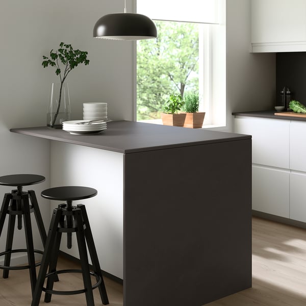 Modern kitchen with large dark grey YTTERVÄGGA waterfall countertop, two black bar stools, black pendant lamp, white cabinets, and a large window.