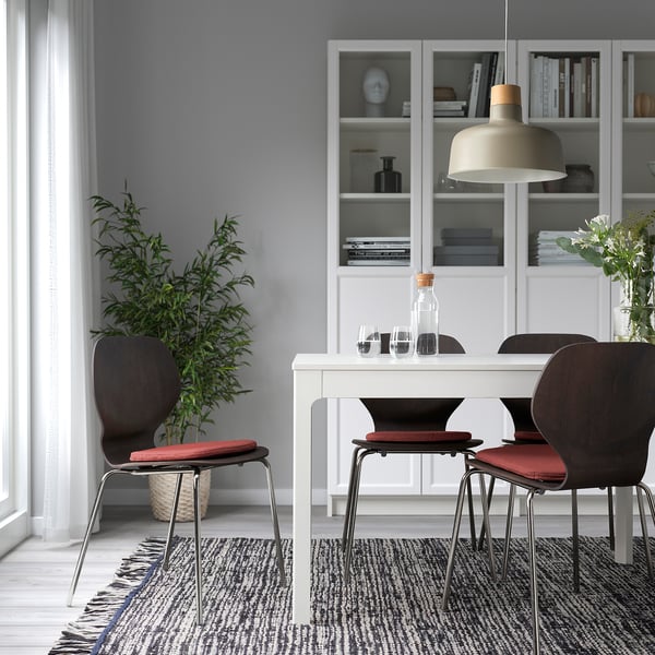 Modern dining area with red chair pads on four chairs around a white table. Houseplant, patterned rug, bookshelf, and pendant lamp.