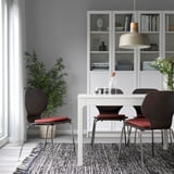 Modern dining area with red chair pads on four chairs around a white table. Houseplant, patterned rug, bookshelf, and pendant lamp.