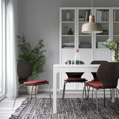 Modern dining area with red chair pads on four chairs around a white table. Houseplant, patterned rug, bookshelf, and pendant lamp.