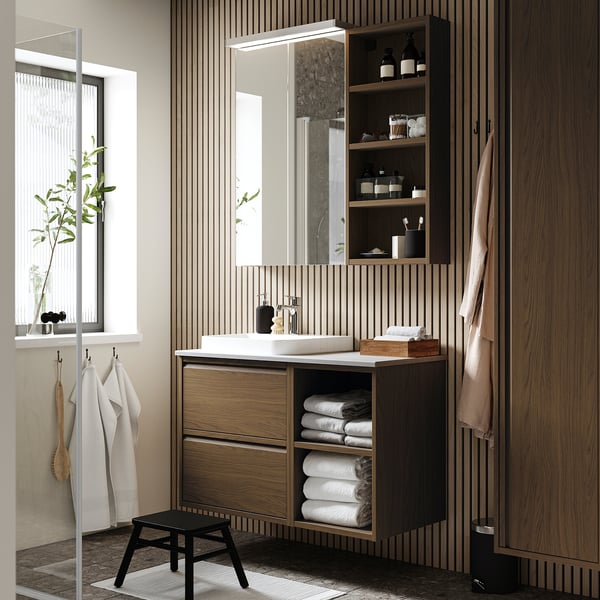 Modern bathroom with wooden cabinet & marble basin, featuring open & closed storage, towels, and plants.