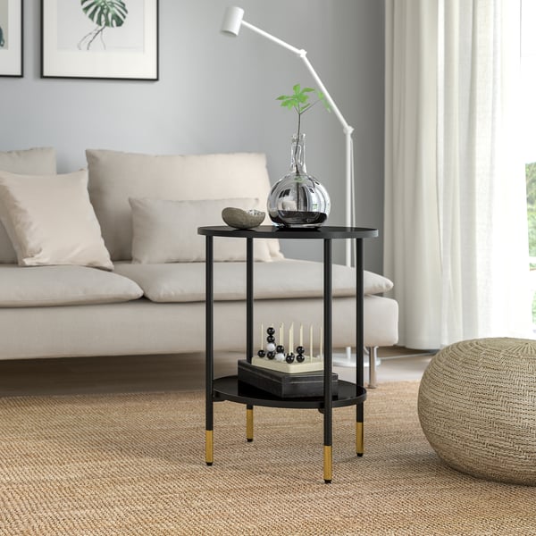 Modern black metal side table with gold accents and glass shelves, beside sofa with vase, plant, and candles.